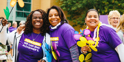 Three women with dark and medium-colored skin smile while wearing purple and holding colorful flowers at a Walk to End Alzheimer's event.