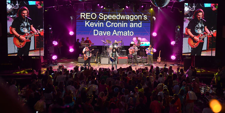 An audience watches a band perform onstage. REO Speedwagon's Kevin Cronin and Dave Amato is shown on a screen.