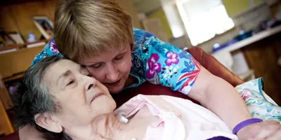 Caregiver embracing an older adult receiving medical care at home, reflecting emotional support and treatment in Alzheimer’s dementia.