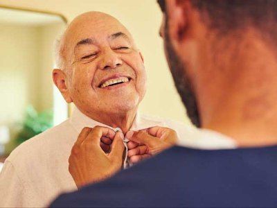 Caregiver assists smiling elderly man in respite car with the buttons of his shirt