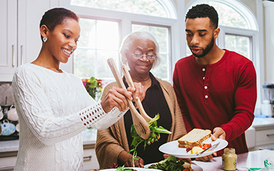 Black family eating together, with a woman putting salad on a plate that a man is holding for an older woman,