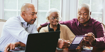An older Black woman and two younger Black men using a computer and tablet and discussing what they see